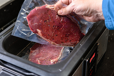 A person places a sirloin steak inside a vacuum sealed bag into a sous vide water bath to cook at 55C. A person places a sirloin steak inside a vacuum sealed bag into a sous vide water bath to cook at 55C.