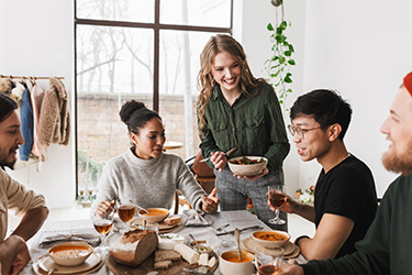 Cheerful woman holding bowl with salad in hand happily spending time with colleagues. Group of attractive international friends sitting at the table full of food having lunch together in cafe